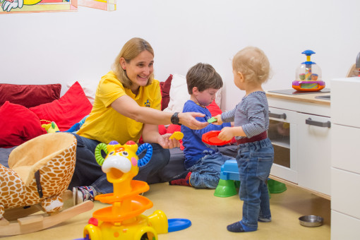 Eine Betreuerin nimmt ein Spielzeug von einem Kleinkind entgegen und lacht 